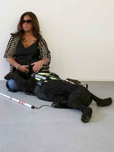 visually impaired woman with glasses sitting on floor with black labradoodle guide dog